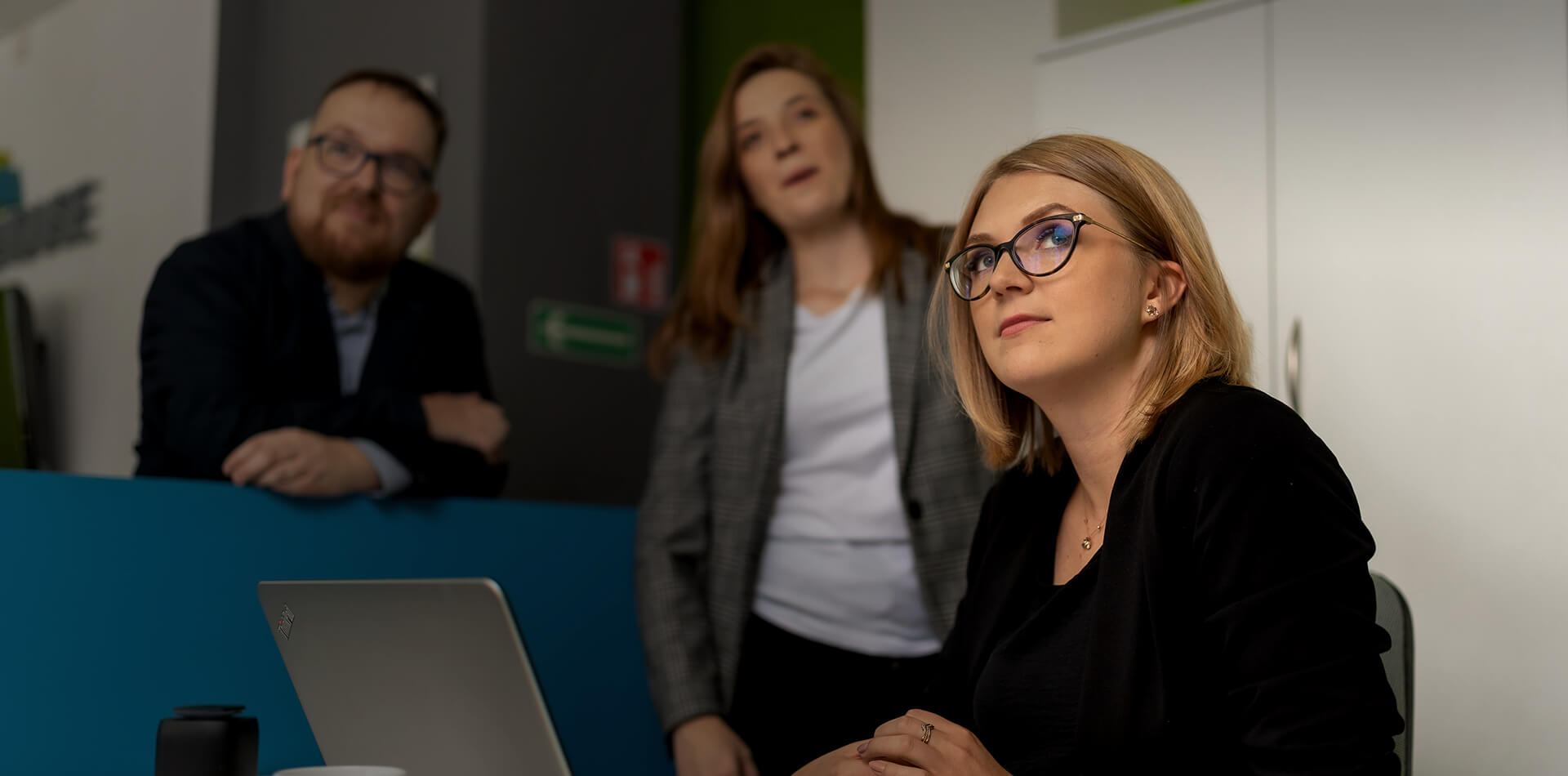 Na zdjęciu widzimy trzy osoby, które znajdują się w biurze. Kobieta siedząca przy biurku o blond, krótkich włosach, w okularach, uważnie patrzy w górę, a przed nią stoi otwarty laptop. Za nią stoi kobieta o ciemnych, dłuższych włosach, ubrana w marynarkę w kratę. Ona również patrzy w górę. W tle po lewej stronie znajduje się mężczyzna w okularach i ciemnej marynarce, który opiera się o blat i patrzy tam gdzie kobiety. 