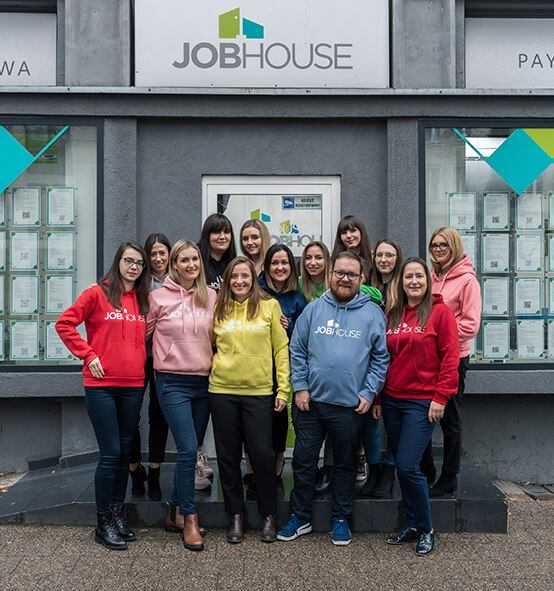 Na zdjęciu widać grupę uśmiechniętych osób stojących przed budynkiem z napisem “JOBHOUSE”. Osoby te mają na sobie kolorowe bluzy z napisem ''JOBHOUSE''. 