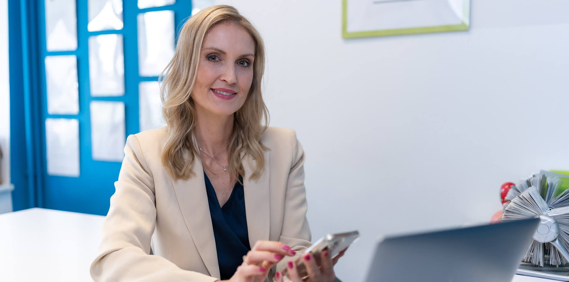 Na obrazie znajduje się uśmiechnięta Natalia Bogdan z blond włosami, siedząca przy biurku. Ubrana jest w elegancką, jasną marynarkę i ciemną bluzkę. Uśmiecha się delikatnie, spoglądając w stronę kamery. W dłoni trzyma smartfon, a na biurku przed nią znajduje się laptop. W tle widoczne są niebieskie drzwi.