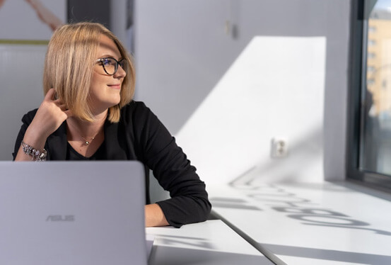 Na zdjęciu widzimy uśmiechniętą kobietę w okularach i krótki, blond włosach, która znajduje się biurze. Kobieta ubrana jest na czarno i siedzi przy biurku z laptopem. Głowa kobiety skierowana jest w kierunku okna po lewej stronie. Na jej twarz padają promienie słoneczne.  W tle widać króluje biel.